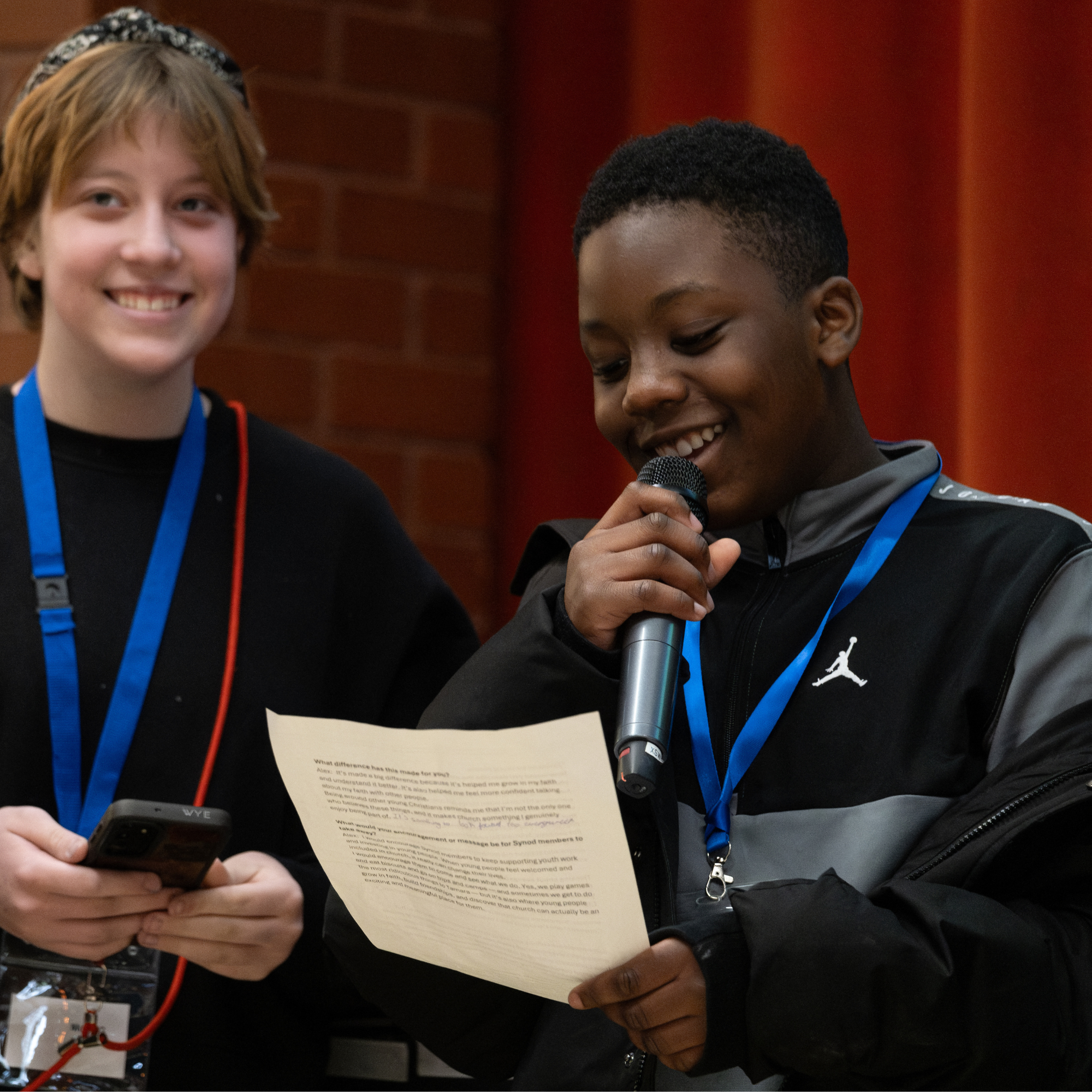 A teenage boy holds a microphone and script as he speaks to a room 