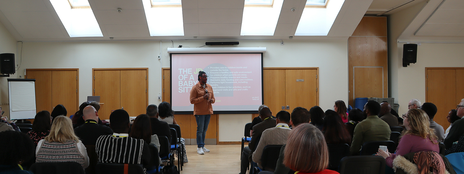 A speaker talks to a power point presentation in front of a room of people on chairs