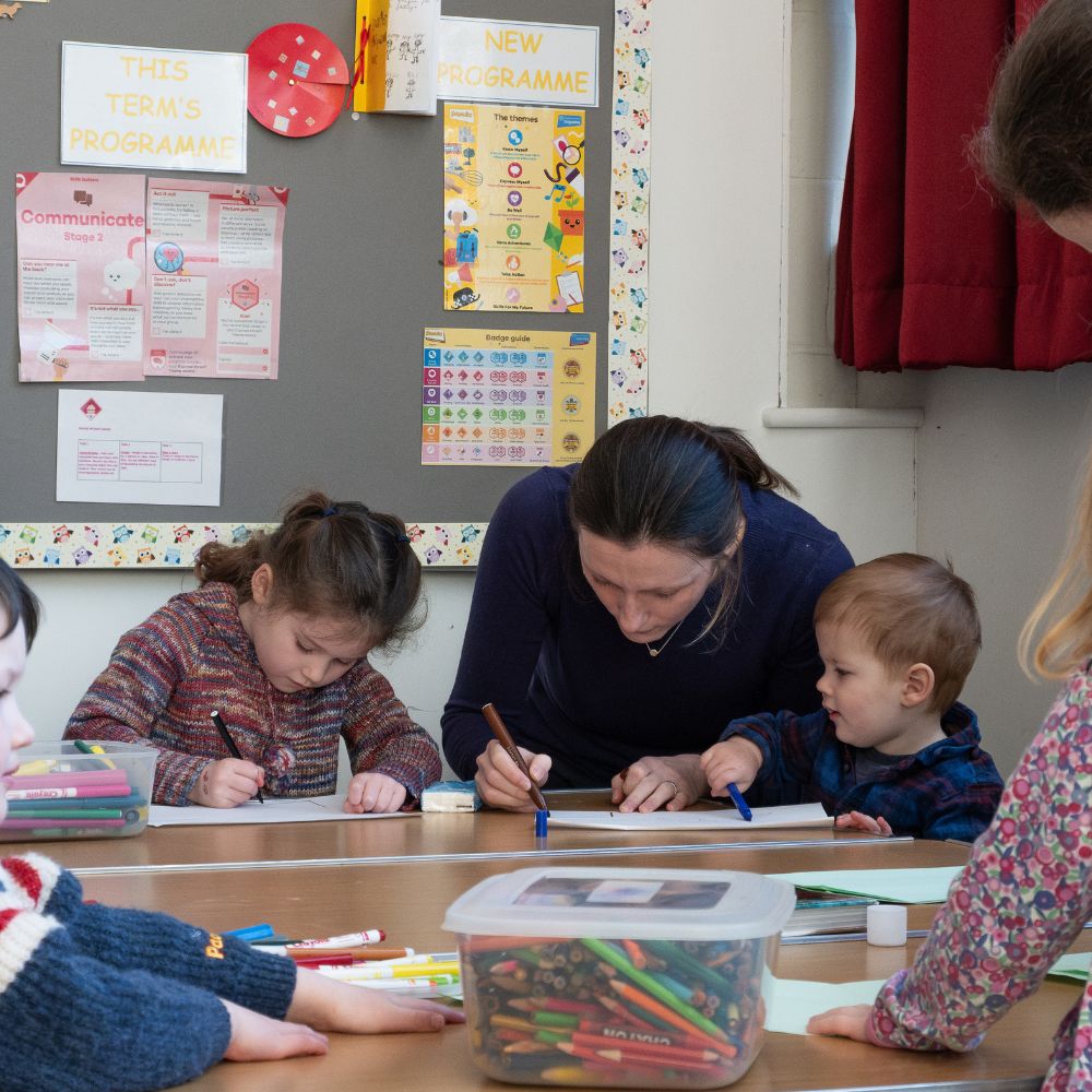 A woman sits at a table surrounded by children doing colouring