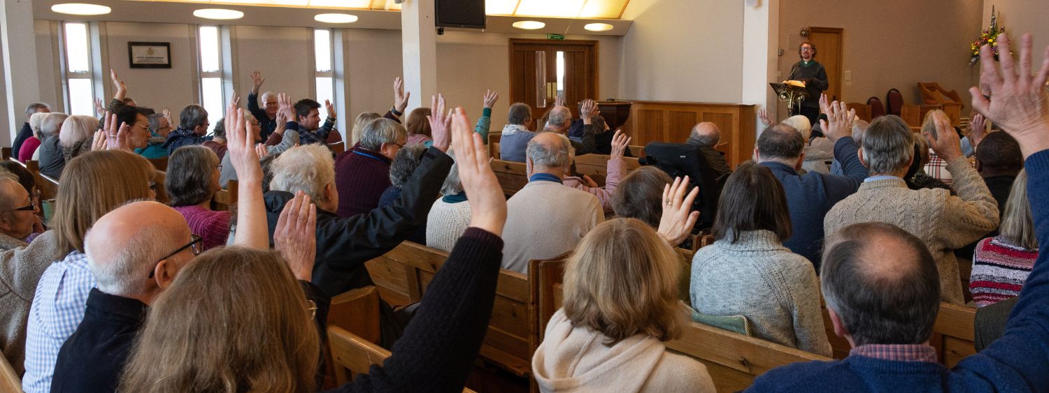 A church congregation facing away from the camera towards the vicar raise their hands