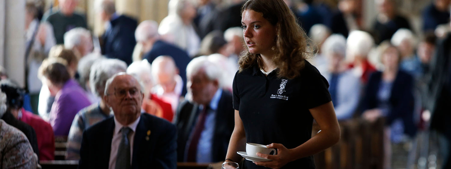 A young woman walks through a busy church holding a mug
