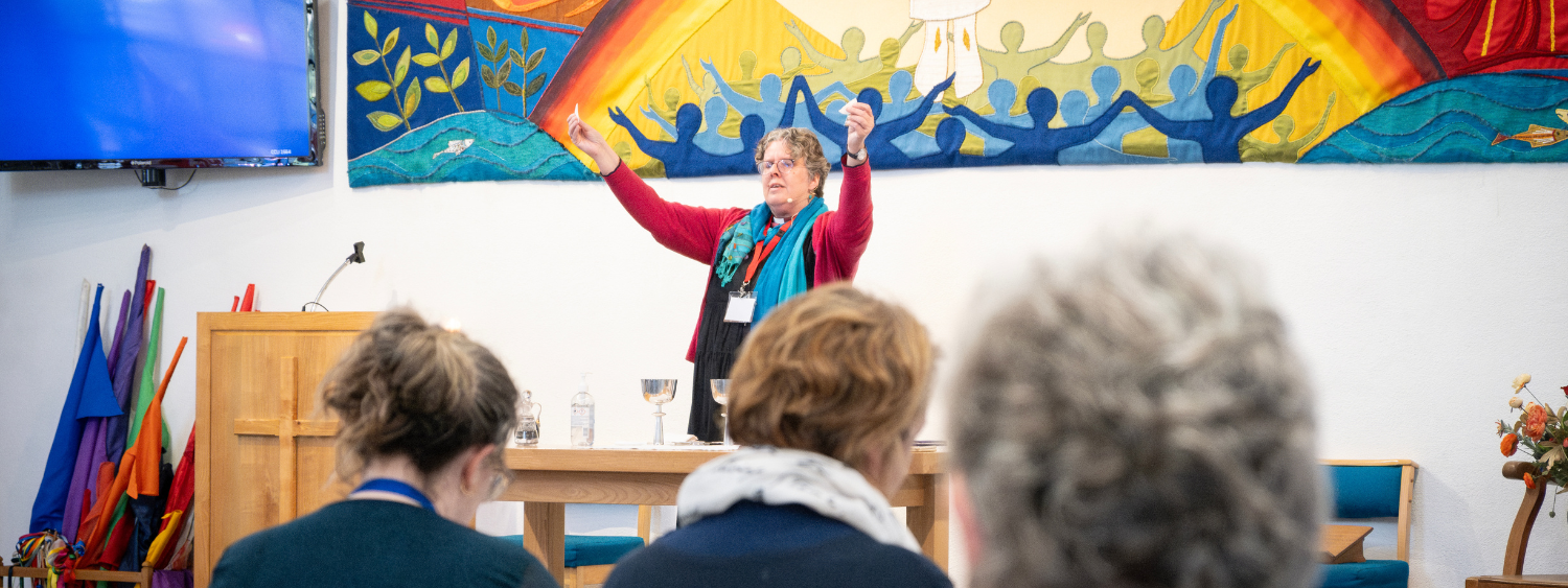 Bishop Mary leads communion, raising bread above her head, in front of a vibrant art piece