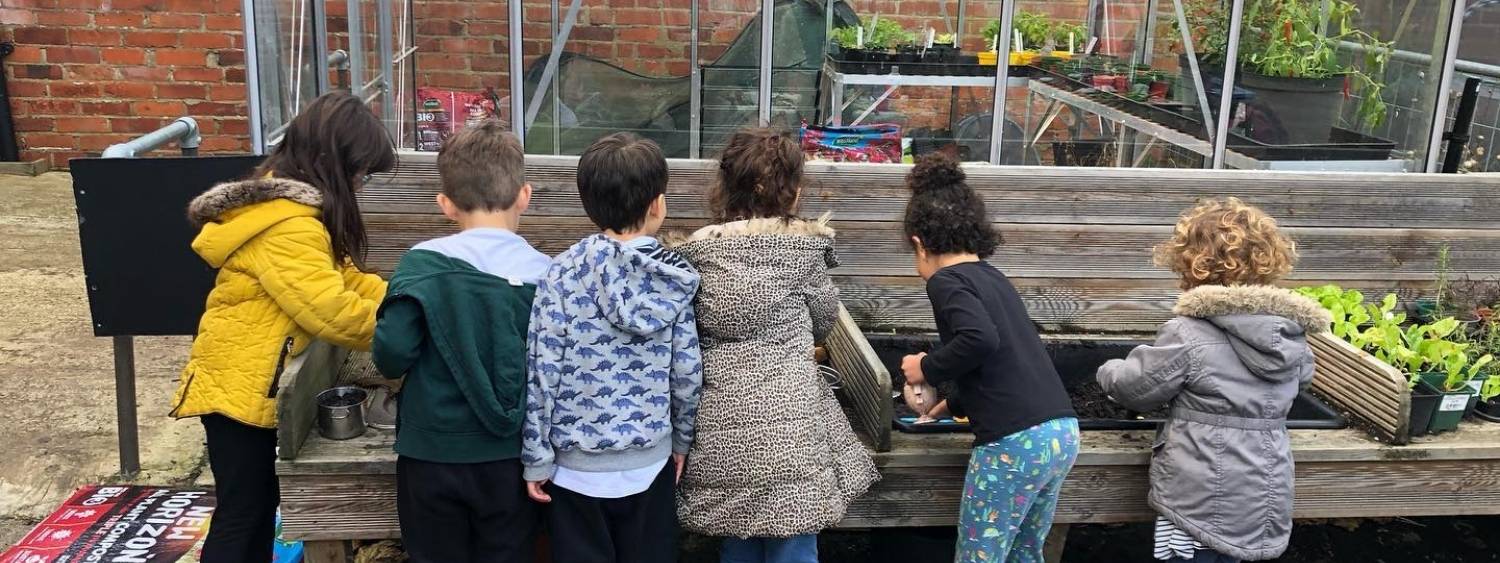 Young children standing in a line in colourful coats playing in a large garden box with mud