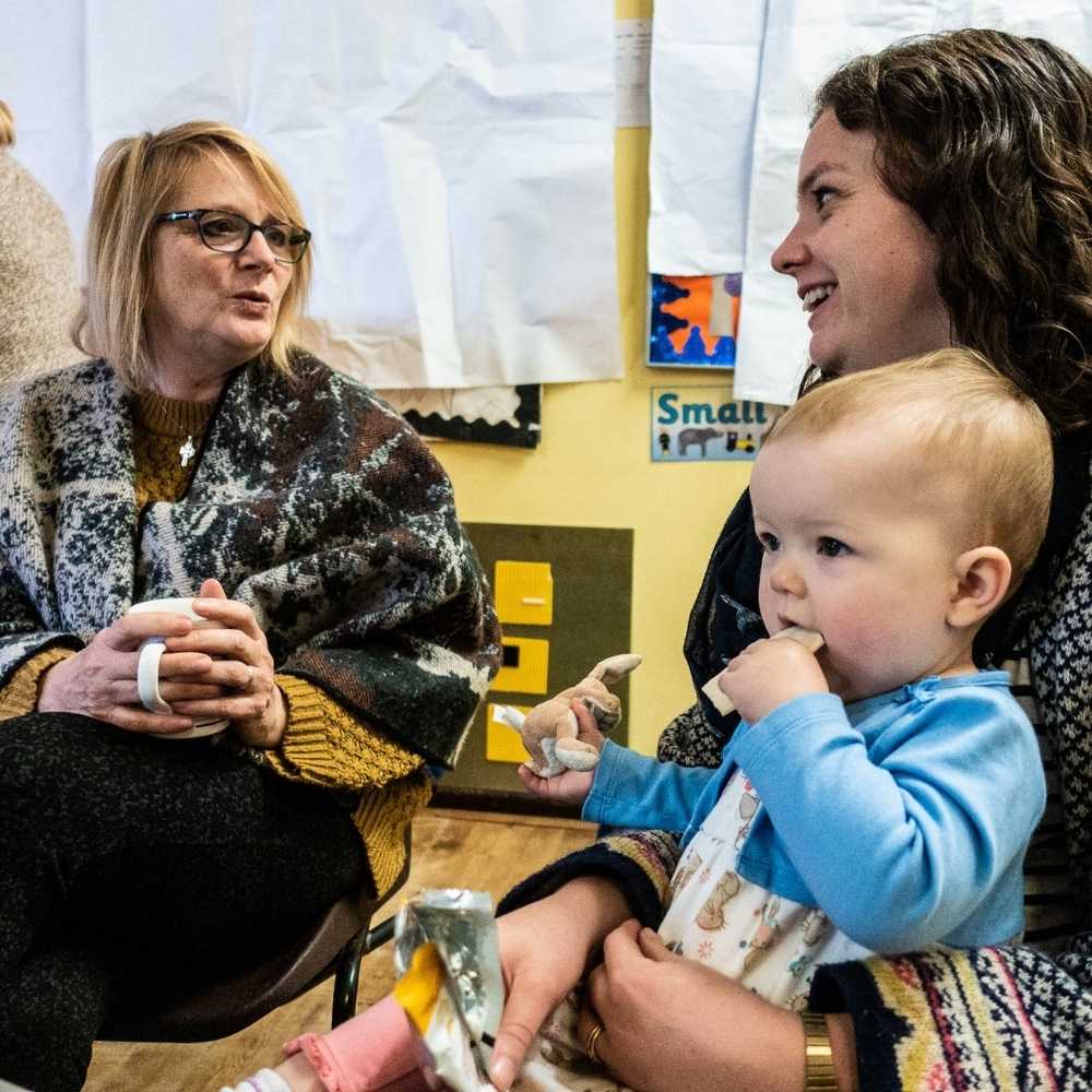 Two women sitting talking and one is holding a toddler who has a hand in their mouth