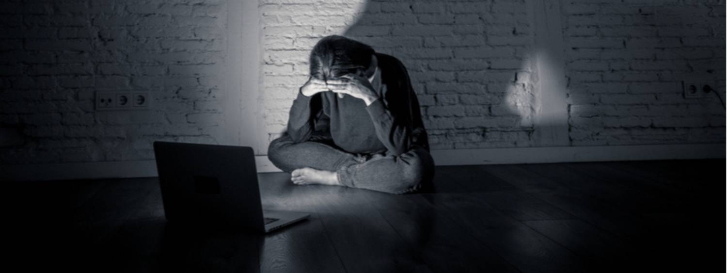 Black and white photograph, young girl sitting cross legged on the floor with head in hands and facing an open laptop