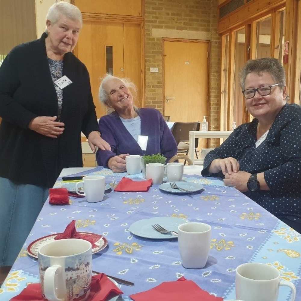 Three women smiling, one stood next to table and two sat at the table with tea and cakfe