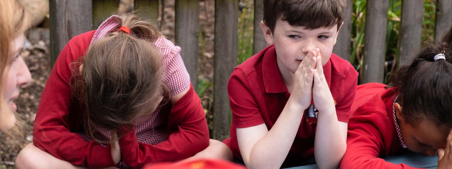 School children sit on a bench outside with their hands together in prayer. Two have their heads bowed while one's eyes are open
