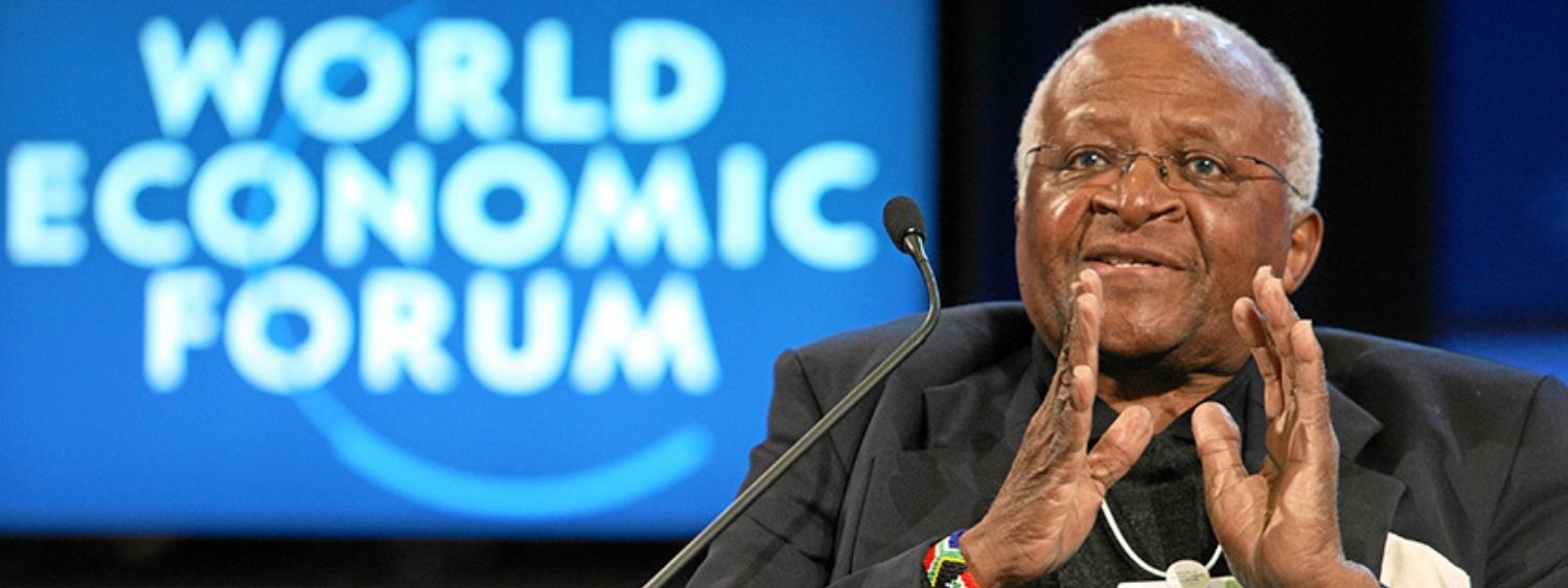 Archbishop Desmond Tutu talking with hands held up and standing in front of a World Economic Forum sign