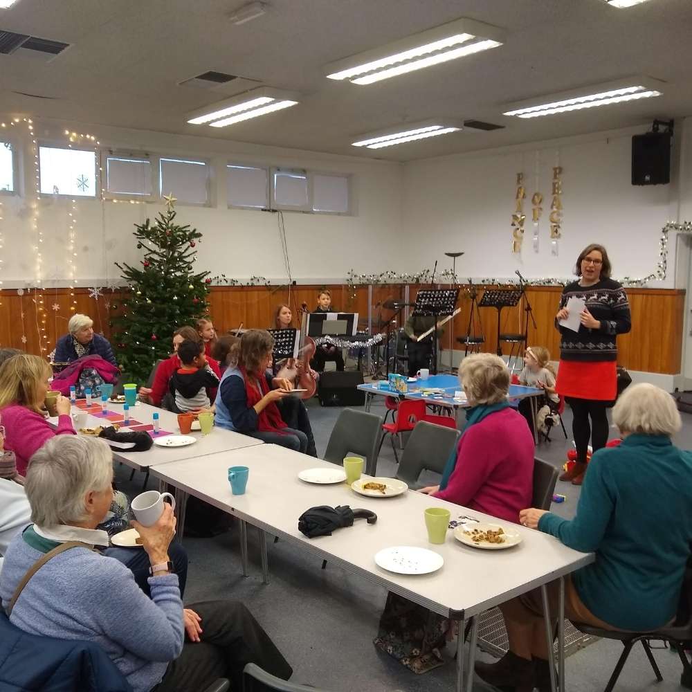 Helen Reid stands in front of a group of older and younger generation people in a church hall sitting around long tables