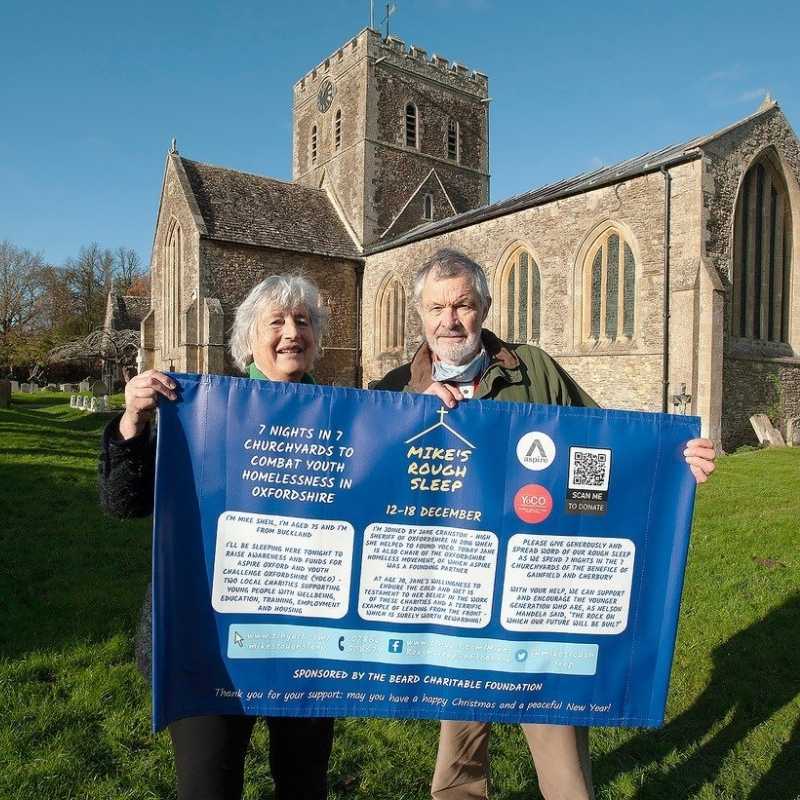 Mike Sheil and Jane Cranston standing outside Oxfordshire Church holding up a blue fundraising banner