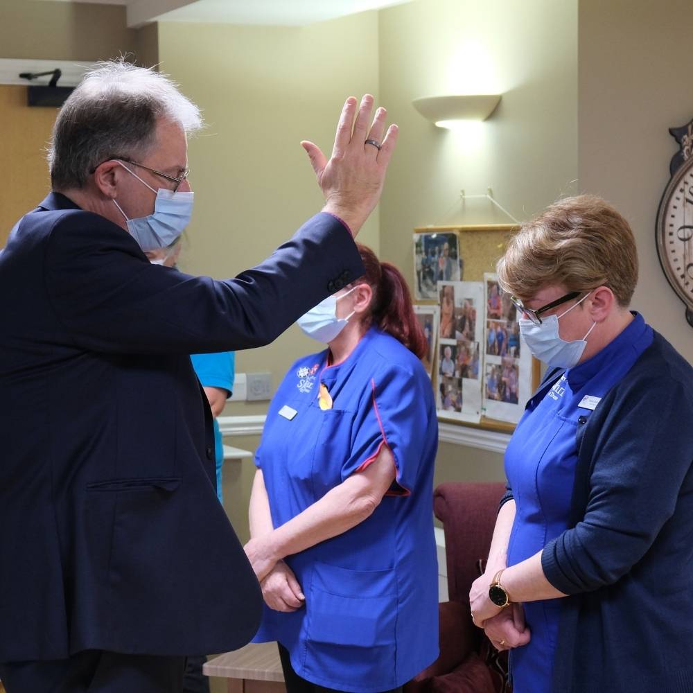 Bishop Steven standing giving a blessing to a female care home worker with his hand raised above her head.