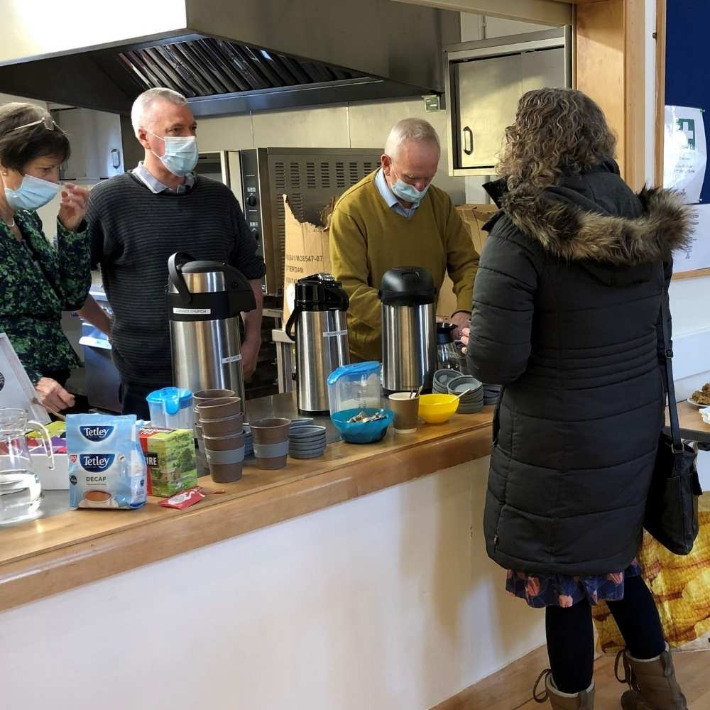 Three volunteers standing behind a counter serving hot drinks to a woman wearing a black coat.