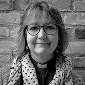 Black and white headshot of the Revd Judith Sumner, standing in front of a brick wall smiling at the camera