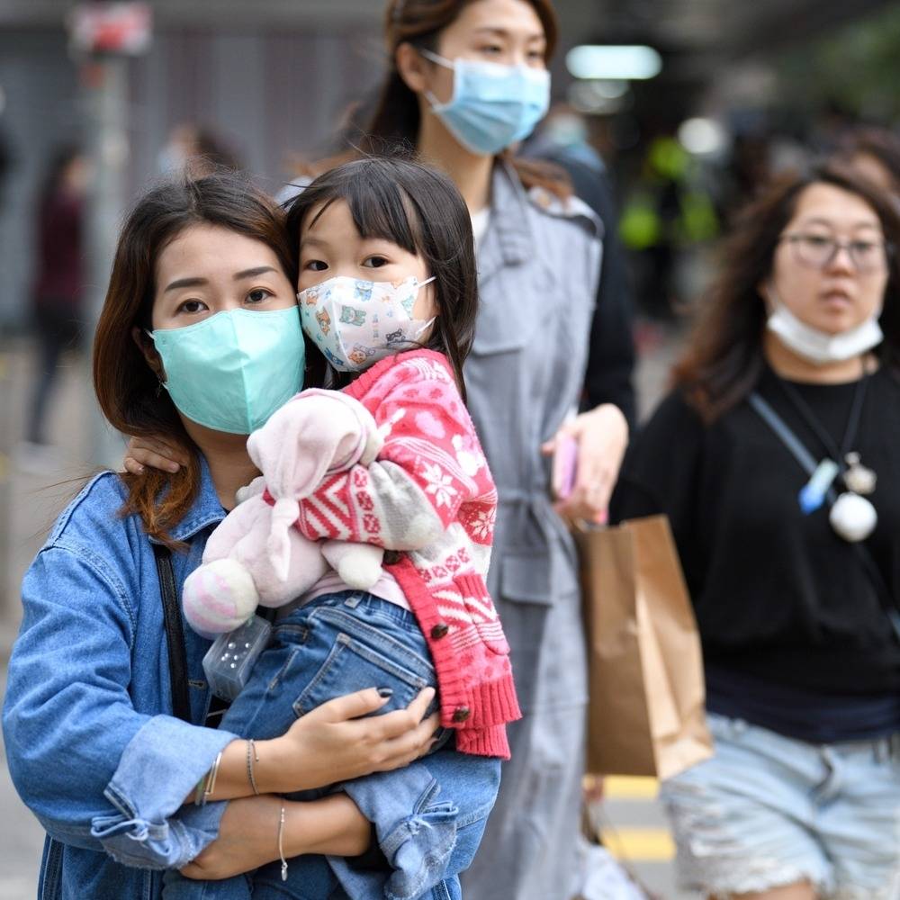 A Hong Kong mother carries her daughter, a toddler with a pink soft toy, both are wearing masks and walking down a street..