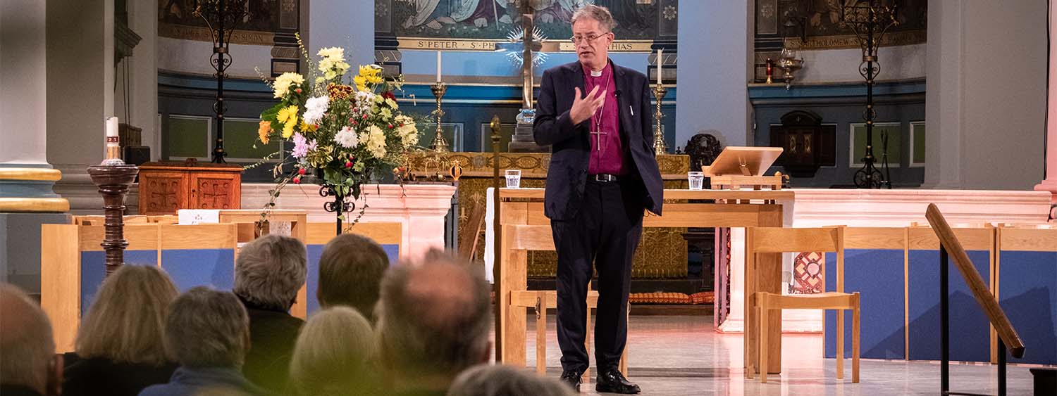 Bishop Steven stands on the stage at St Mary's Church, Banbury, looking at the congregation as he preaches. A large arrangement of flowers and golden religious art is in the background.