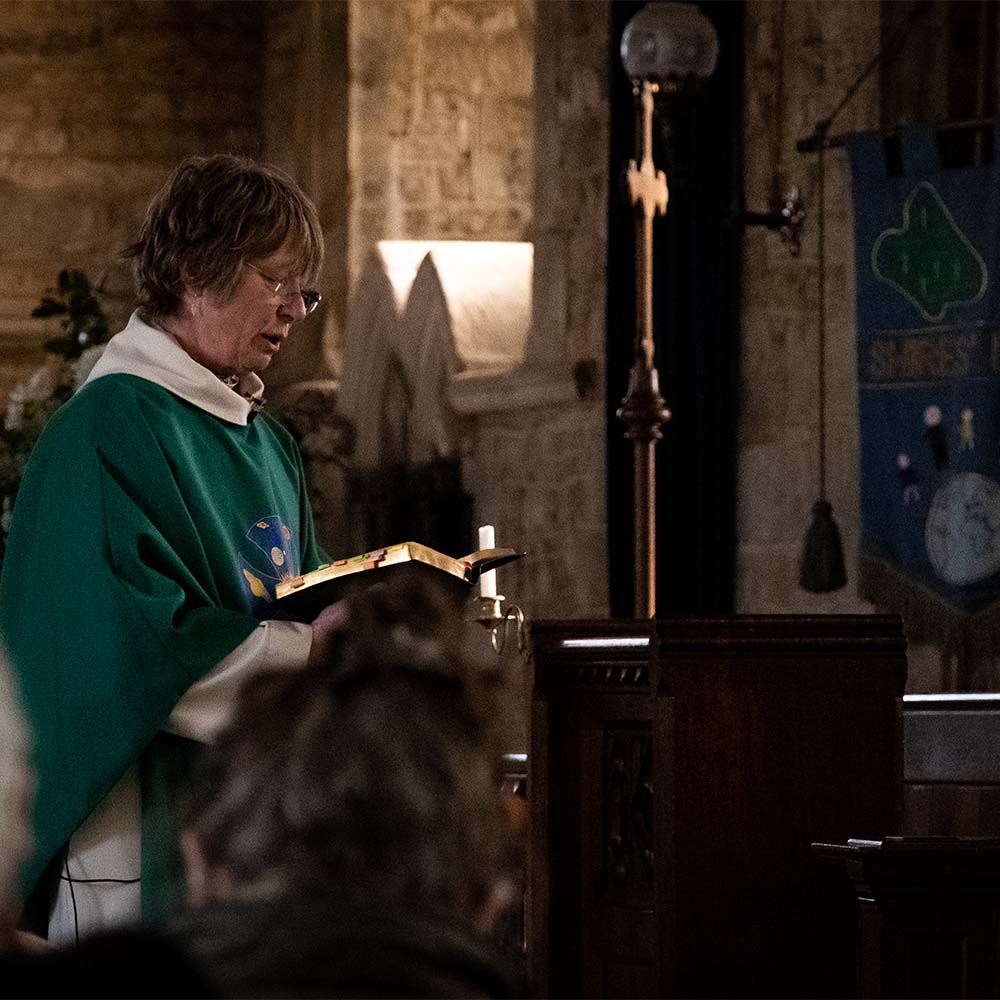The Revd Hillary Campbell holds the Gospel as she reads from it to the church congregation. The Revd Hillary wears a green robe, and the book has coloured notes marking various pages.