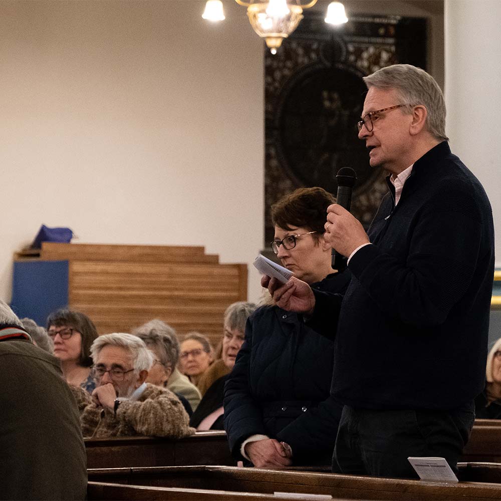 A man stands in a church pew speaking into a microphone and holding prompt cards. The rest of the congregation look forward towards Bishop Steven, off camera.