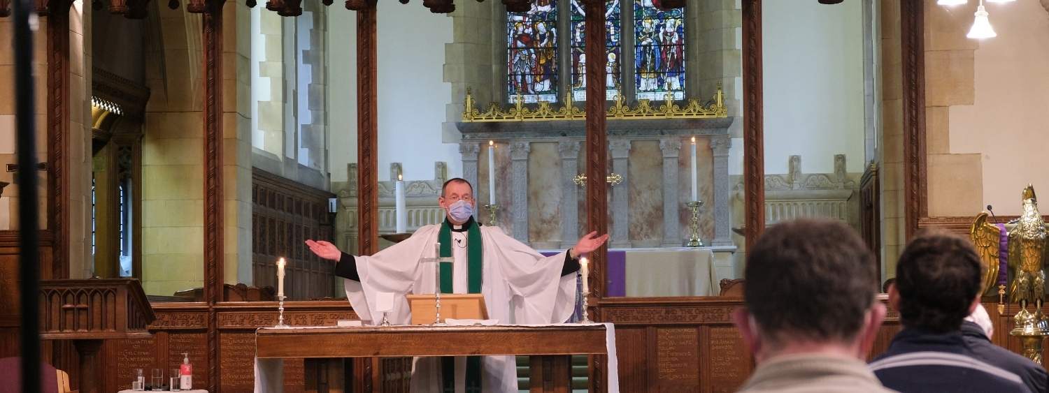 The Revd Steve stands with his arms outstretched at the front of church wearing white robes and a dark green stole. He is looking at congregation members.