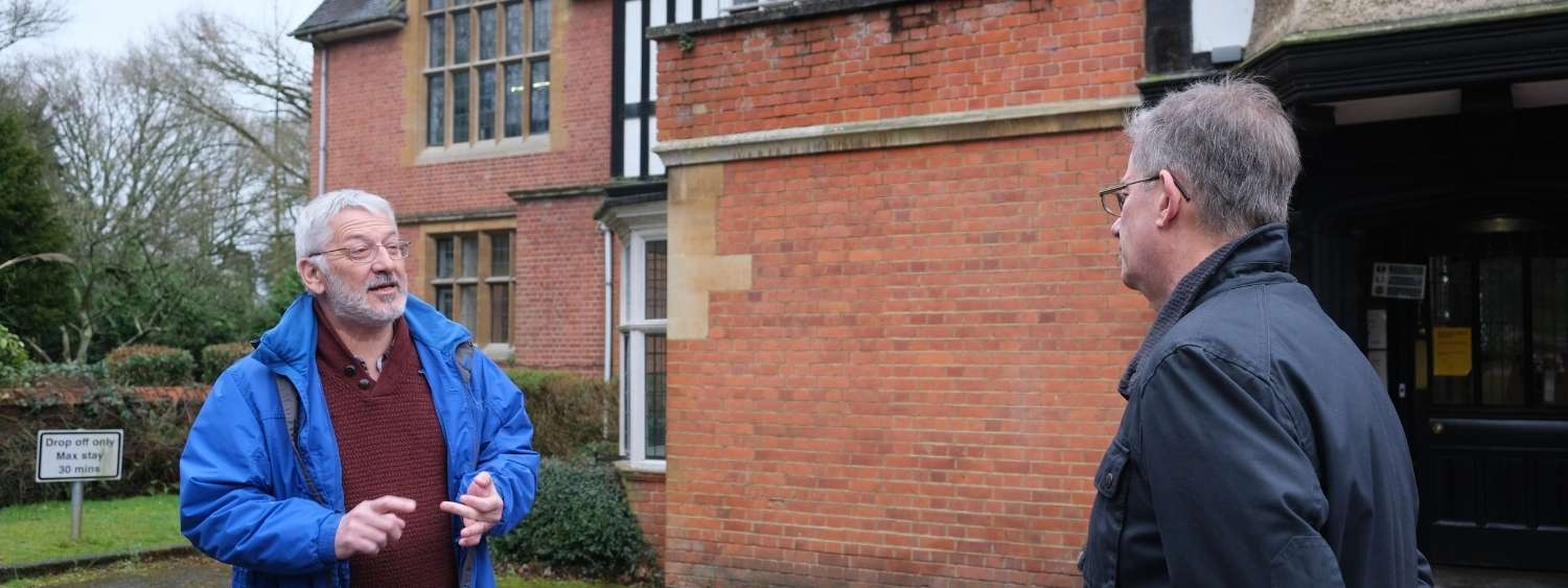 Chris Wood and Bishop Steven standing outside the briick entrance to Yeldall Manor as Chris shares the history of Yeldall with Bishop Steven. Chris is wearing a blue raincoat and Bishop Steven a dark navy jacket.