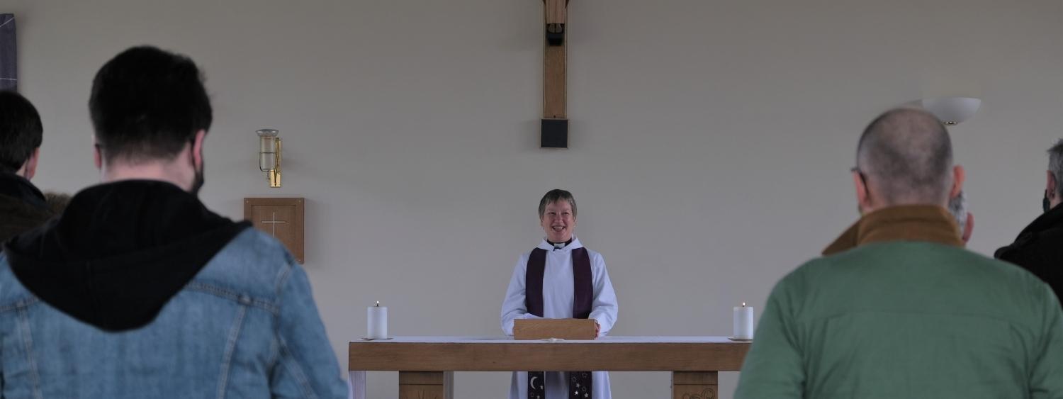 The Revd Dr Liz Ratcliffe is standing smiling at the standing congregation at the Altar table at the front of St Nic's Earley church wearing white robes and a purple stole.