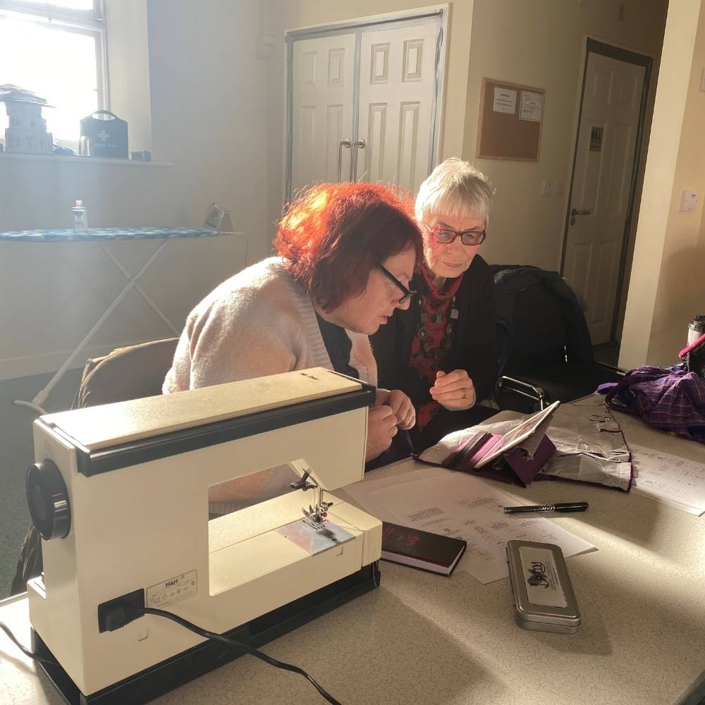 Two women sitting at a table with a sewing machine and looking at an iPad.