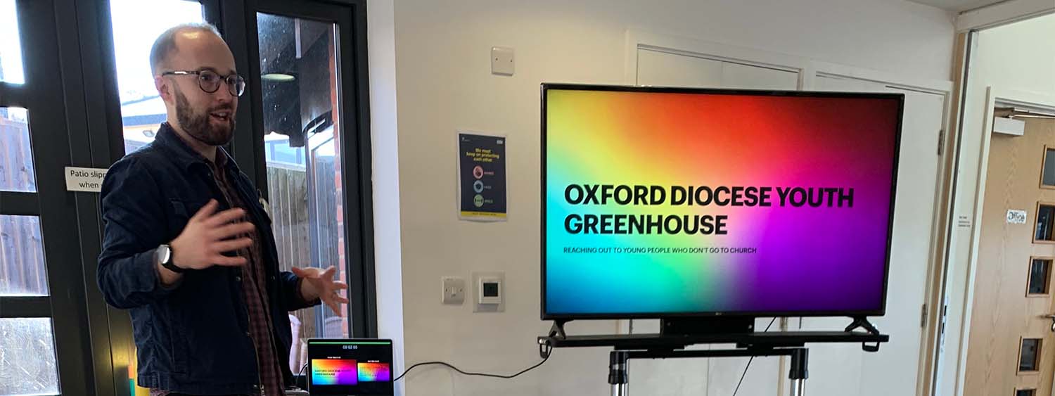 Olly Shaw, Youth Greenhouse convenor, presents to a group of people out of shot. He stands next to a screen with a colourful display that reads 