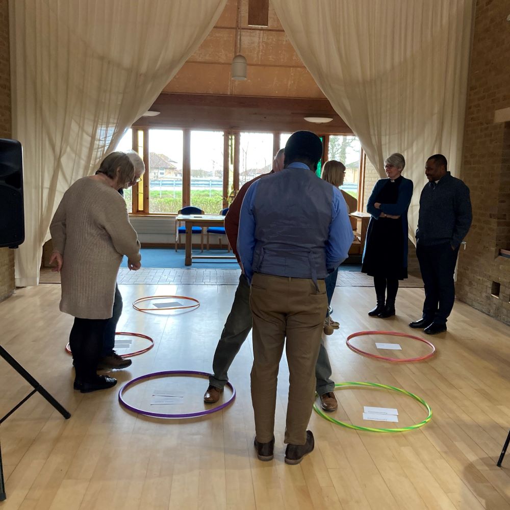 A group of people stand in a circle in a church hall. Om the floor is a circle of large hoops, each with a piece of paper in the middle. People stand at or in the hoop(s) that best represent where they are in the Loving First Cycle