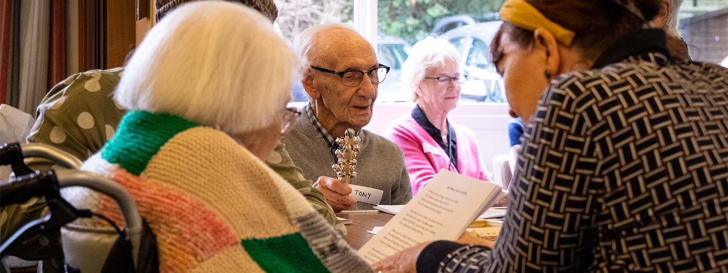 A group of people sit around a table looking at a music sheet. An elderly man shakes a jingle stick,