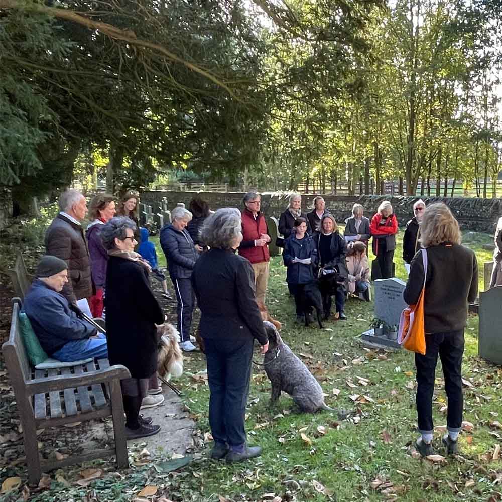 A large group of people of all ages stand and sit in a churchyard, dressed warmly, as they listen to a woman facing away from the camera reading prayers. Some people have brought their dogs