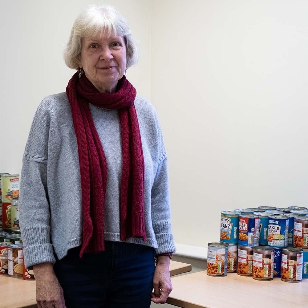 Jo Anderson stands in a room next to a table. On the table is a small pyramid of tins of baked beans. Jo is smiling and wearing a long knitted scarf, a woolly jumper and small dangly earrings