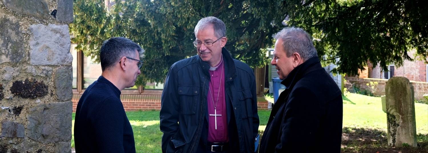 Bishop Steven standing talking to two male clergy in Warfield Parish Church yard in front of a large tree and headstones.