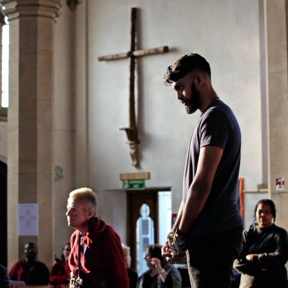 Man with a beard standing with hands tied together with rope and a crucifix is on the wall in the background.