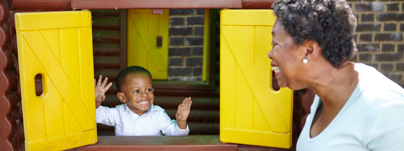A small boy grins as he pokes his head through the window of a wendy house. Blurred in the foreground, an adult smiles back at him