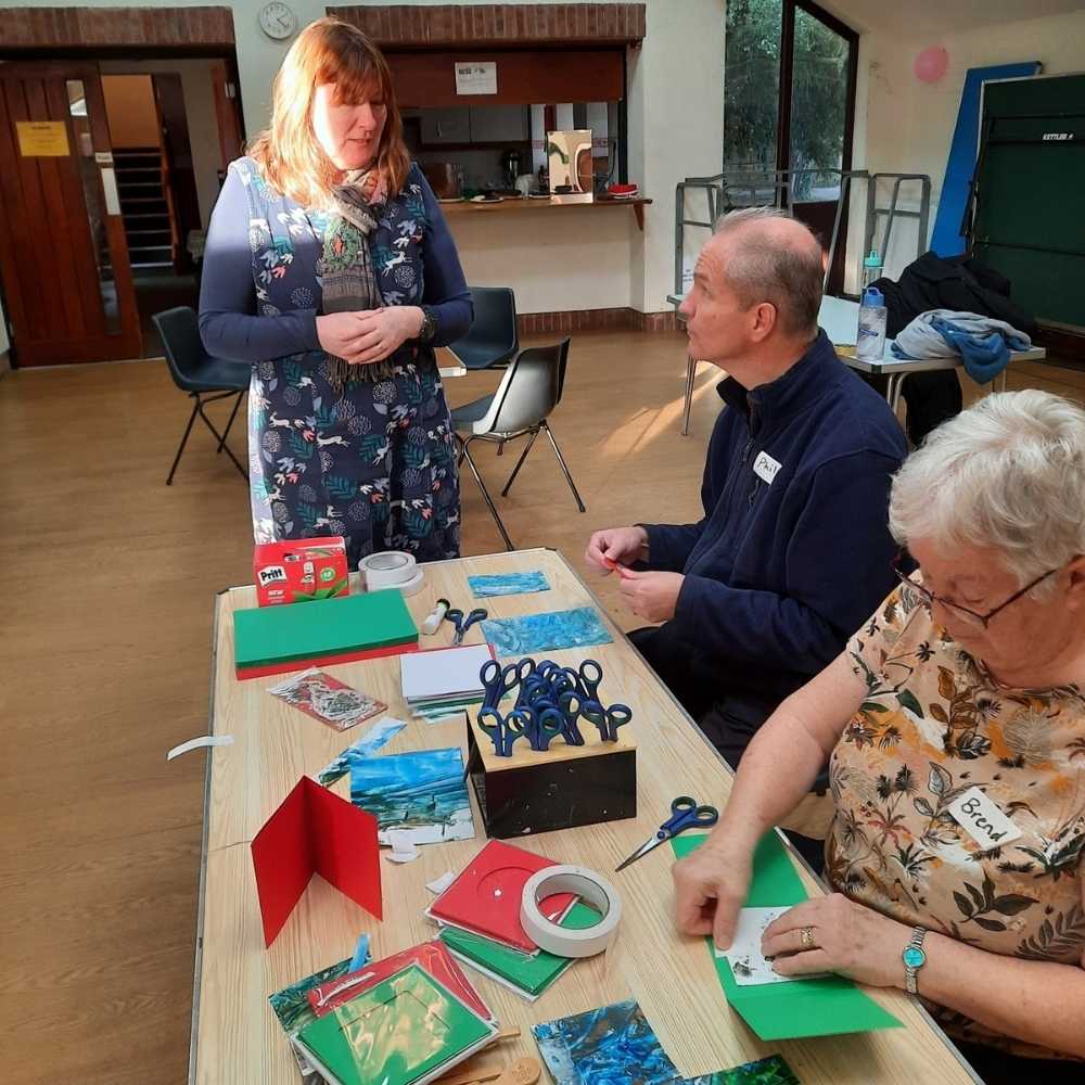 An older man and woman sitting at a table with craft items, the man is looking up and talking to a woman standing next to the table.