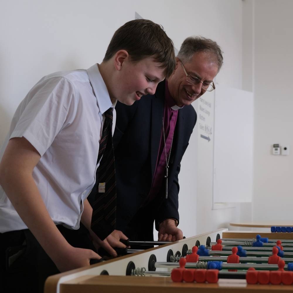 Bishop Steven and a young teenage boy who is wearing a school shirt and tie playing table football and smiling.