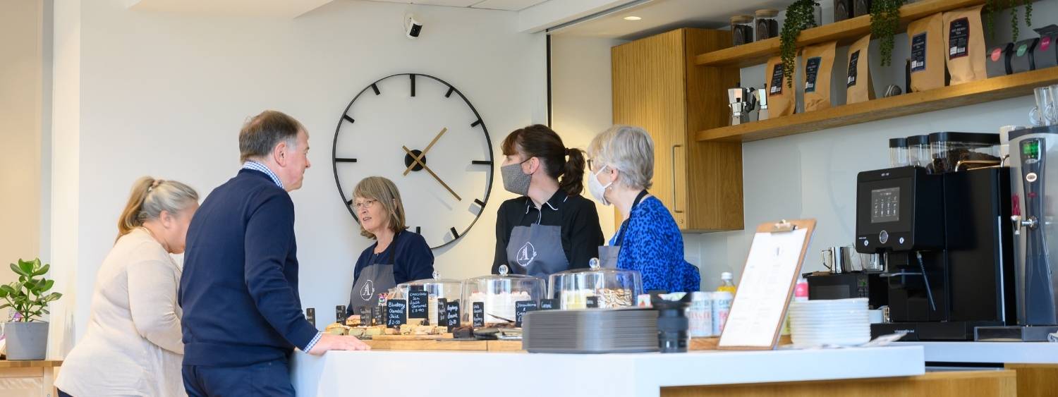 a cafe bar with 3 female staff members stood behind serving 2 customers, cakes are displayed on the counter, coffee bags on shelves in the background and there is a large clock on the wall.
