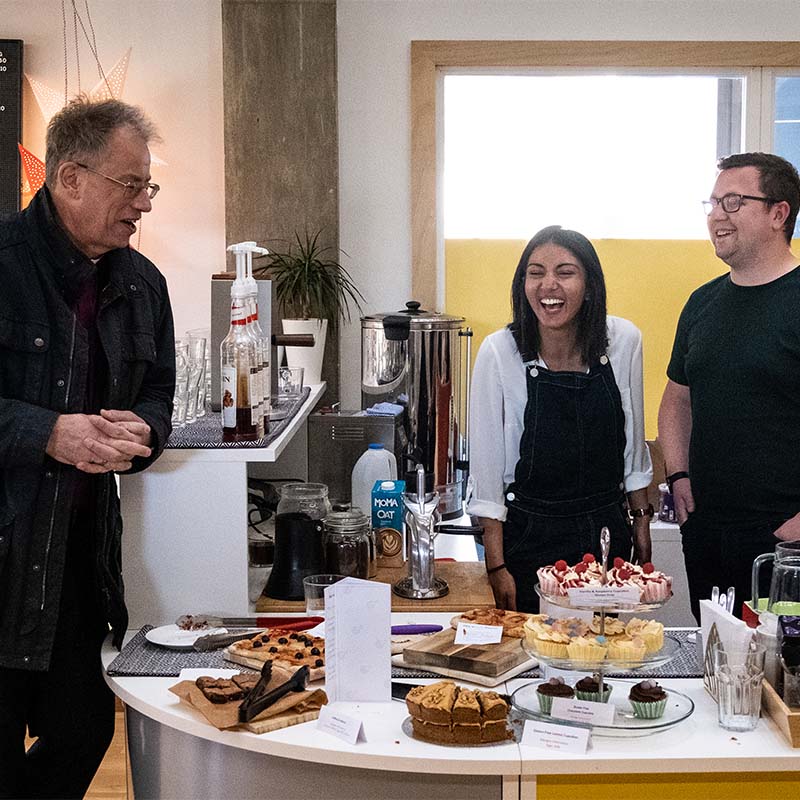 Bishop Steven laughs along with two cafe workers who stand behind a counter full of cakes