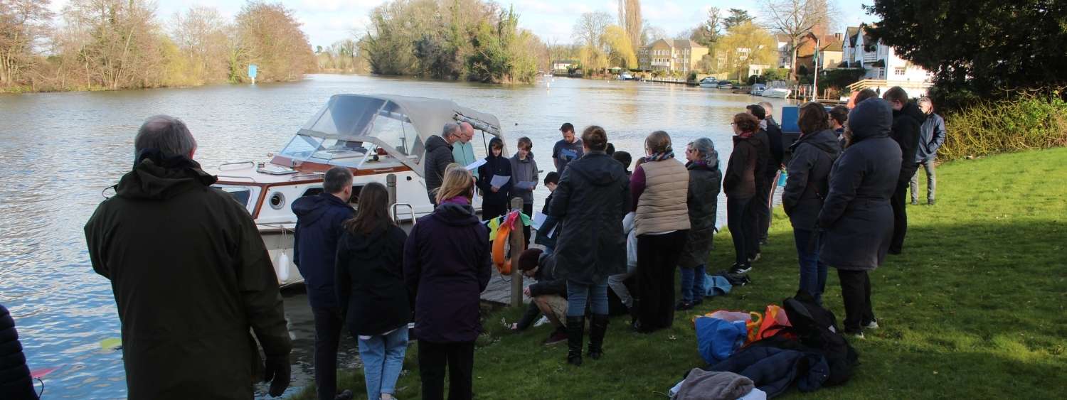 A group of people are standing on a grass bank next to the River Thames in Bray, there is a boat on the water and two young teenage boys are preparing to be baptised.