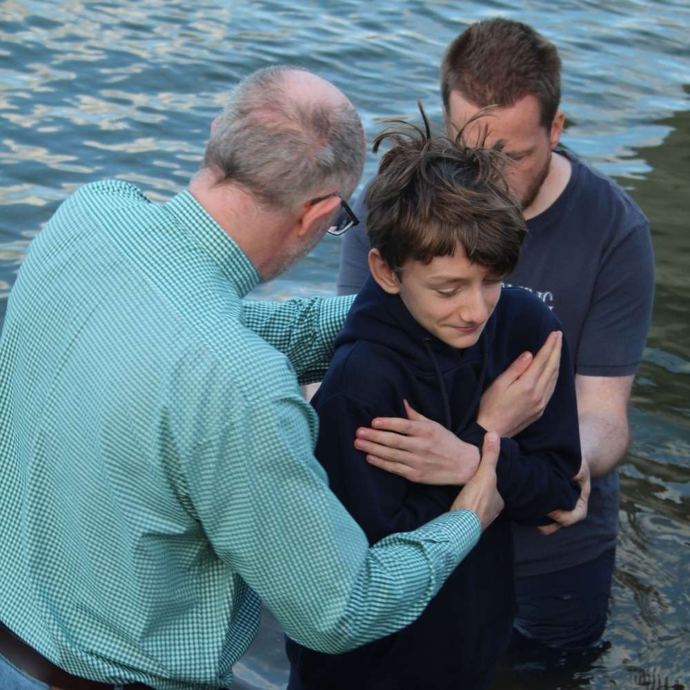 A young teenage boy is standing with his hands crossed over his chest at waist height in the river Thames with two adult men holding him ready to guide him under the water.