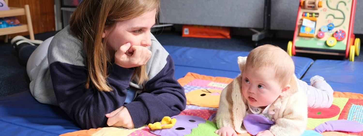 a mother and her baby girl lying on their front on a soft blue play mat, there are toys in the background.