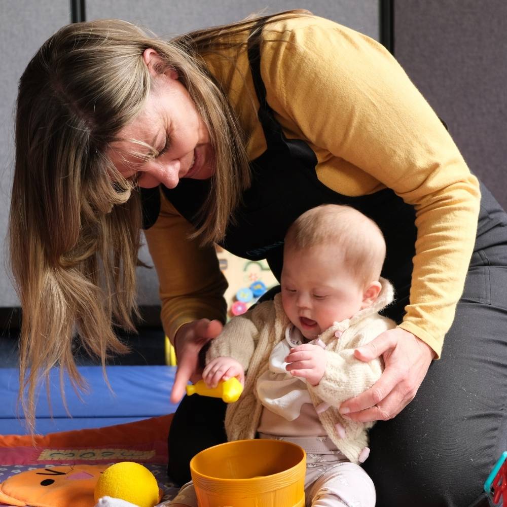 Estelle is kneeling on a soft blue play mat and holding a baby girl in front of her on the floor whilst she plays with toys.