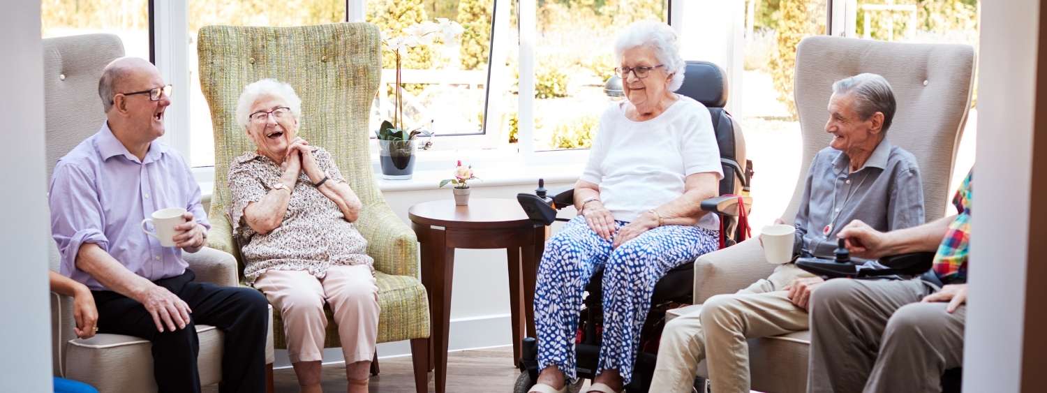 Four elderly men and women sitting in chairs at a care home laughing and drinking hot drinks