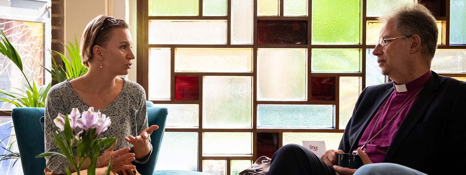 Bishop Steven sits in a cafe talking to Yulia Johnson. Behind them is a stained glass wall.