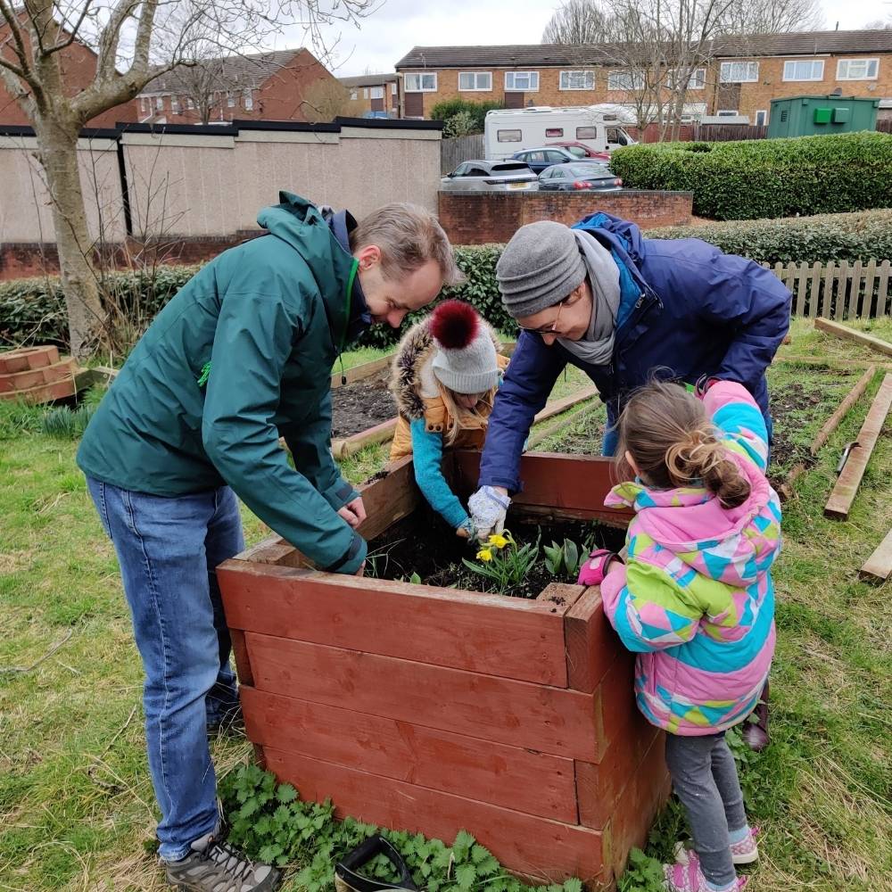 two adults and two young children are digging in a large wooden box for plants.