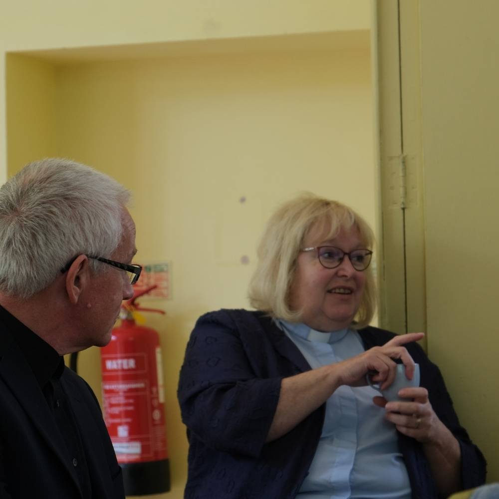 Female clergy holding a blue cup and speaking, a male clergy person is sitting listening to her.