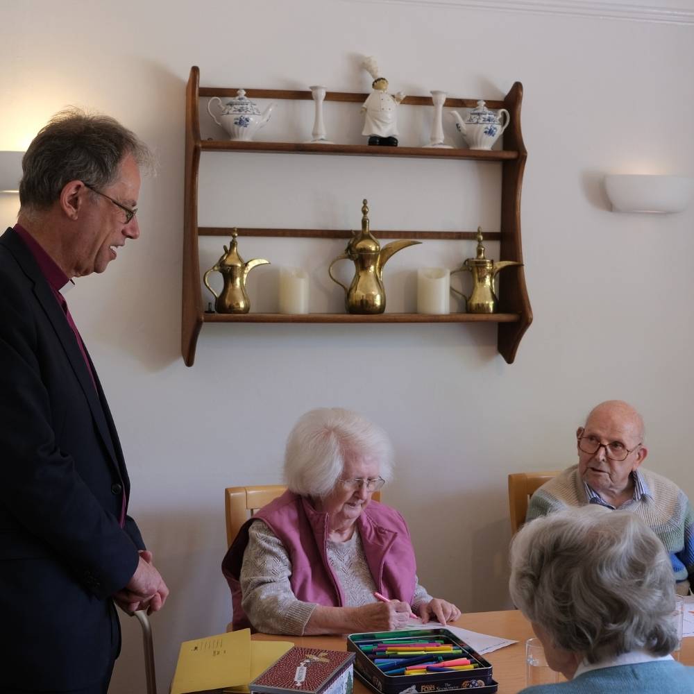 Bishop Steven standing talking to residents at Coombe House seated at a table.