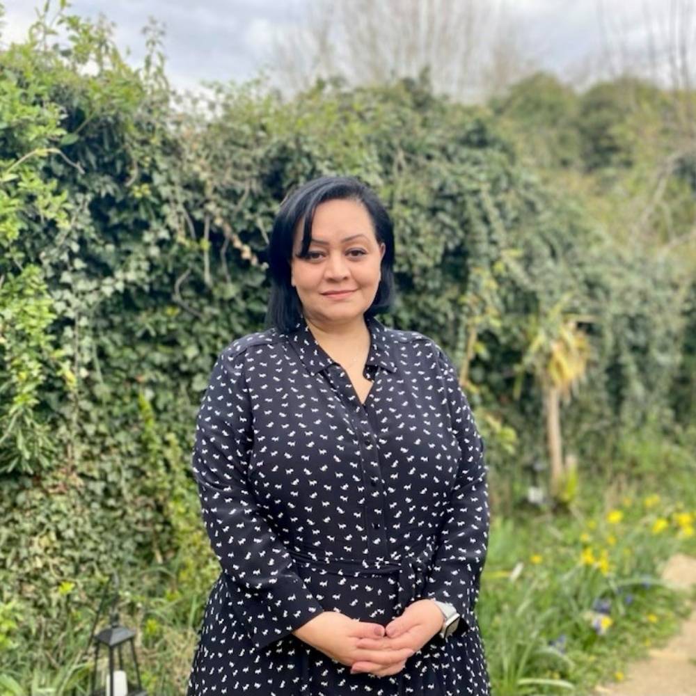 Azita is standing smiling outside in front of a hedge and has her hands clasped infront of her