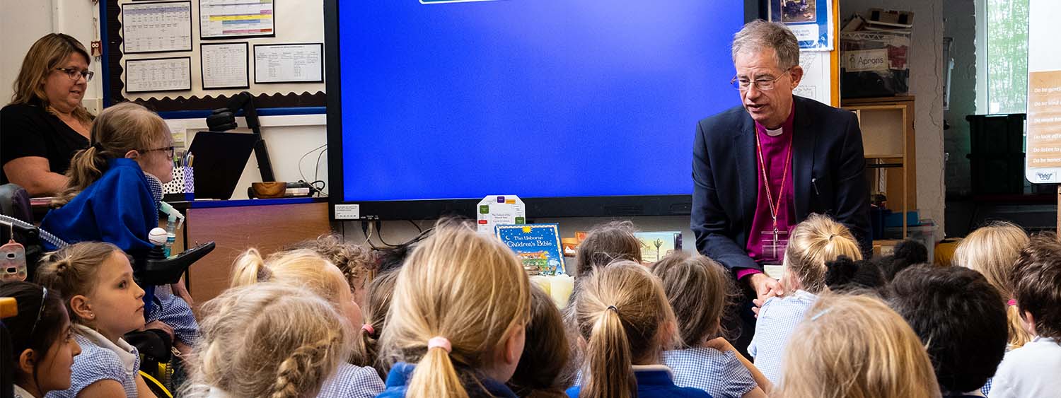 Bishop Steven sits at the front of a classroom, talking to a group of primary school students, who sit on the floor looking up at him