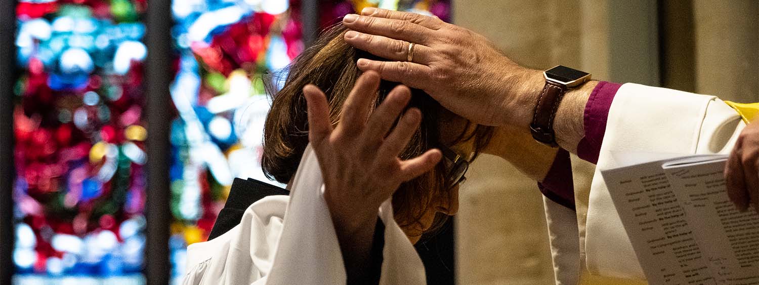 Close up photo of an ordinand being ordained deacon. Her head is bowed and her arms are outstretched. The Bishop of Oxford's hands are placed on her head. In the background is a vibrant stained glass window at Christ Church Cathedral, Oxford