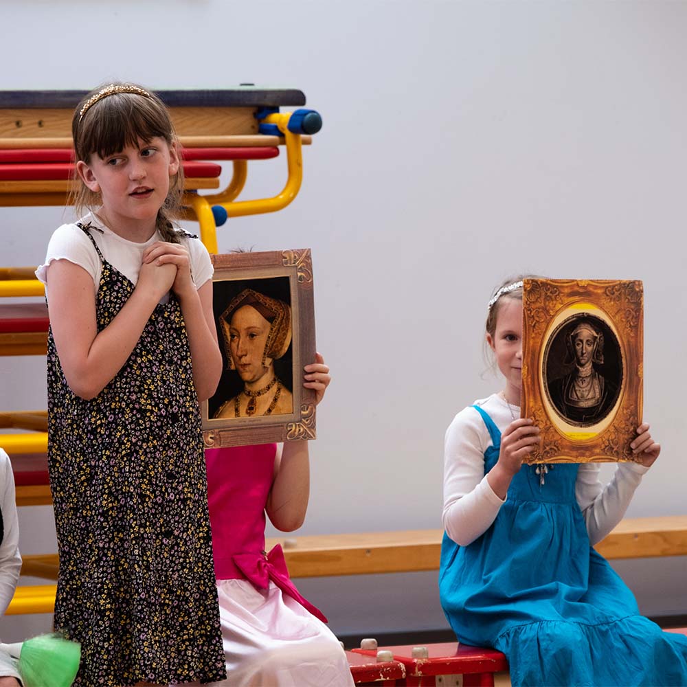 School children perform an assembly about Henry VIII and his wives. A girl stands and delivers her lines. Behind her two children hold portraits of Henry's wives over their faces.
